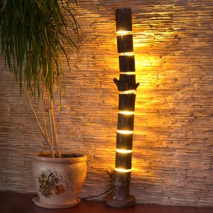 May include: A rustic wooden floor lamp with a unique design. The lamp is made from stacked sections of a tree trunk, with warm light emanating from the spaces between the sections. A potted plant sits to the left.