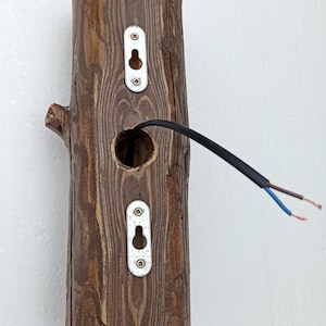 May include: A wooden wall sconce with a hole for a light bulb and two metal plates with keyholes. The sconce is attached to a white wall with black electrical wires.