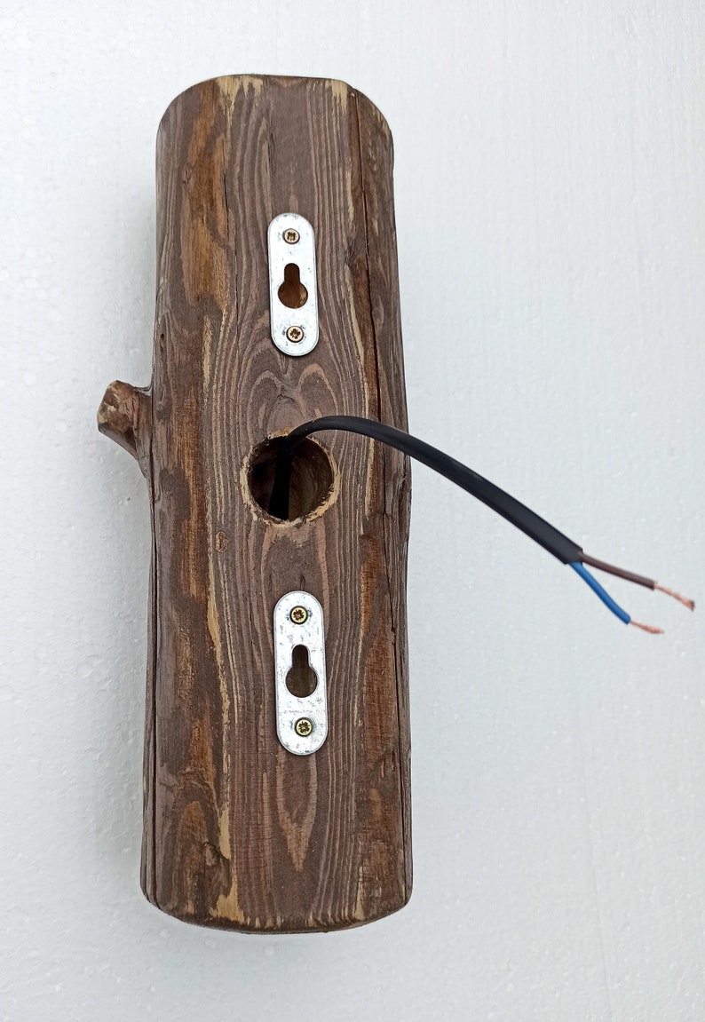 May include: A wooden wall sconce with a hole for a light bulb and two metal plates with keyholes. The sconce is attached to a white wall with black electrical wires.