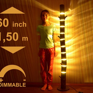 May include: A dimmable, wooden floor lamp, approximately 60 inches tall, with a tree trunk design. The lamp has multiple light sources and a warm glow. The base is round, and the lamp is plugged into an electrical outlet.