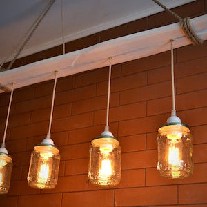 Könnte beinhalten: Ein rustikaler Kronleuchter mit vier Glasbehältern als Leuchten, die an einer weißen Holzplanke mit Seil-Akzenten hängen. Die Behälter haben klares Glas und weiße Porzellansockel mit warmweißen Glühbirnen, die den Raum erhellen.