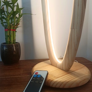 May include: A wooden table lamp with a white LED light and a remote control. The lamp has a unique curved design and a natural wood finish. The remote control has a black and white design with buttons for power, brightness, and color temperature.