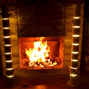 May include: Two wooden floor lamps with a tree trunk design stand on either side of a fireplace. The lamps are made of stacked cylindrical sections that emit a warm, yellow light. A fire burns brightly in the fireplace.