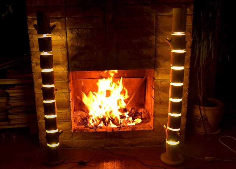 May include: Two wooden floor lamps with a tree trunk design stand on either side of a fireplace. The lamps are made of stacked cylindrical sections that emit a warm, yellow light. A fire burns brightly in the fireplace.