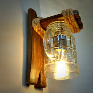 May include: A rustic wall sconce made from wood and rope with a clear glass jar light fixture. The jar has a handle and a light bulb inside.