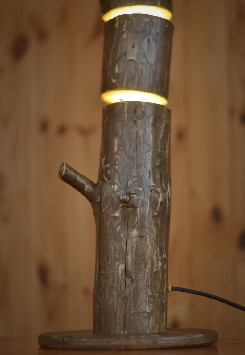 May include: A rustic wooden lamp designed to resemble a tree trunk. The lamp has a natural brown finish with visible wood grain and a small branch. The lamp has three sections that emit a warm, inviting light. A black power cord extends from the base.