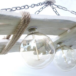 Puede incluir: Una l&aacute;mpara de ara&ntilde;a de madera r&uacute;stica con un acabado blanqueado y detalles de cuerda. La l&aacute;mpara de ara&ntilde;a tiene cuatro bombillas de globo de vidrio transparente.