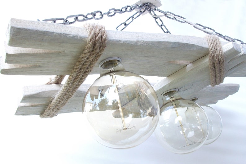 Puede incluir: Una l&aacute;mpara de ara&ntilde;a de madera r&uacute;stica con un acabado blanqueado y detalles de cuerda. La l&aacute;mpara de ara&ntilde;a tiene cuatro bombillas de globo de vidrio transparente.