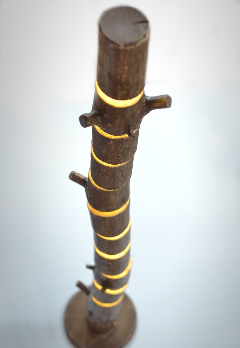May include: A brown, tree-trunk-shaped lamp with a round base. The lamp has several small branches and emits a warm, yellow light from the cut-out sections. The lamp is made of wood and has a natural, rustic appearance.