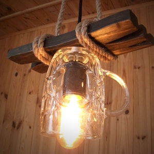Peut inclure: Un luminaire rustique fait d'une chope à bière réutilisée, suspendue à une poutre en bois avec une corde. La chope est en verre transparent avec une surface texturée et une poignée. Une ampoule éclaire l'intérieur de la chope.