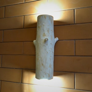 May include: A white wooden wall sconce shaped like a tree trunk with a branch extending out. The sconce is mounted on a brick wall and is illuminated by a light bulb inside.