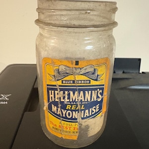 Może przedstawiać: Antyczny, przezroczysty szklany słoik z wyblakłą etykietą. Etykieta zawiera niebieską wstążkę i napis "Hellmann's Real Mayonnaise". Słoik jest lekko zakurzony, co sugeruje wiek. Widoczny jest napis "Blue Ribbon".