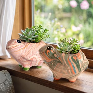 May include: Two ceramic animal-shaped planters with marbled finishes. The left planter features pink and white swirls with green feet, while the right planter has orange, blue, and grey swirls with orange feet. Both contain green succulent plants.