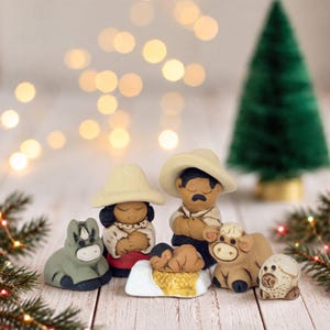 May include: A ceramic nativity set featuring a central figure with a wide-brimmed hat, a figure with a smaller hat, a swaddled infant on a white base, a gray donkey, a brown cow, and a small white sheep, all with hand-painted details.