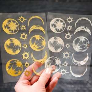 May include: Two sheets of clear adhesive paper with gold and silver foil stickers. The stickers feature various celestial designs, including moons, stars, and suns.
