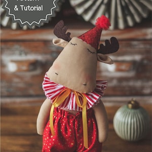Pode incluir: Um brinquedo de pelúcia de alce vestindo um colar listrado vermelho e branco, um macacão estampado com estrelas vermelhas e douradas e um chapéu de festa vermelho com glitter e um pompom vermelho. O alce tem chifres marrons e os olhos fechados.