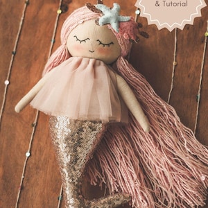 May include: A handmade pink and gold mermaid doll with long pink yarn hair and a sparkly gold tail. The doll is wearing a pink tulle skirt and has a starfish on her head. The doll is sitting on a wooden surface.