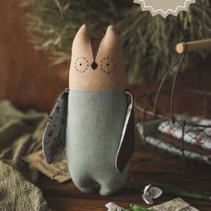 Pode incluir: Um brinquedo de pelúcia de coruja marrom e bege com asas marrons e um corpo estampado em azul e branco. A coruja está sentada em uma superfície de madeira com um livro e flores.