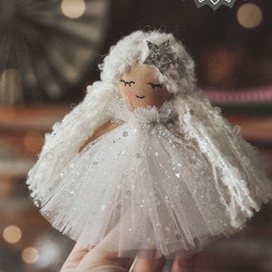 Puede incluir: Una muñeca de ángel de tela blanca con una estrella plateada en la cabeza y una falda de tul blanca con purpurina plateada. La muñeca está siendo sostenida por una mano.