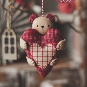 May include: A red and white plaid heart-shaped ornament with a brown teddy bear face and small arms. The ornament is hanging from a red and white string. The text "PDF Pattern & Tutorial" is on a red starburst.