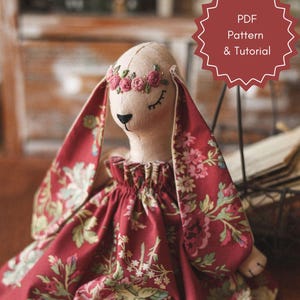 Puede incluir: Una muñeca de conejo de tela hecha a mano con un vestido rojo floral y una corona de flores. La muñeca tiene los ojos cerrados y una expresión dulce. La muñeca está sentada sobre una superficie de madera con un carrete de hilo cerca. Patrón PDF y Tutorial.