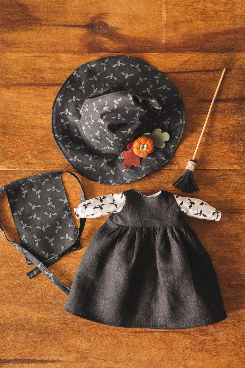 May include: A black witch costume for a doll, including a hat, dress, and a small bag. The hat and bag have a bat print. The dress has a black bodice and a black skirt with a gathered ruffle.
