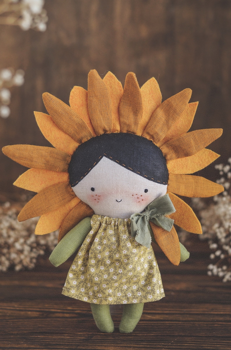 K&ouml;nnte beinhalten: Eine handgefertigte Puppe mit einem Sonnenblumenkopf, einem gr&uuml;nen Kleid mit Blumenmuster und gr&uuml;nen Armen und Beinen. Die Puppe steht auf einer Holzoberfl&auml;che.