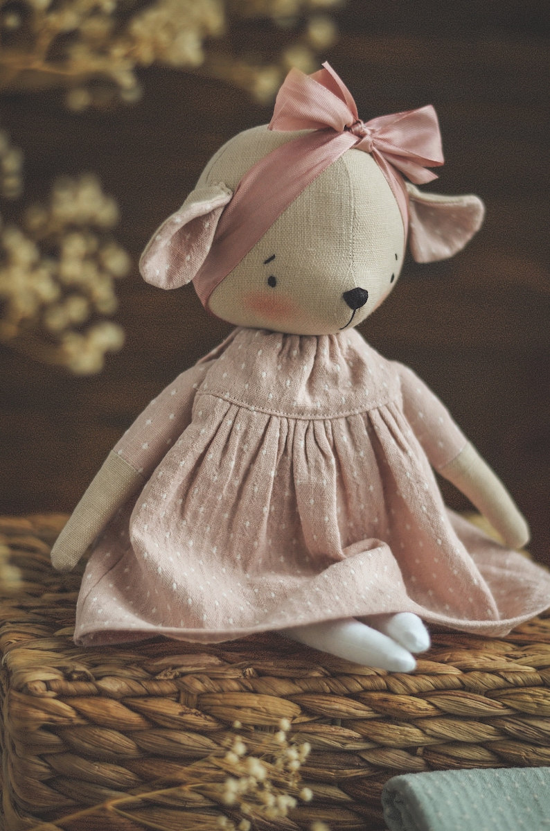 May include: A handmade plush toy in the shape of a deer wearing a pink dress and a pink bow. The toy is sitting on a woven basket.