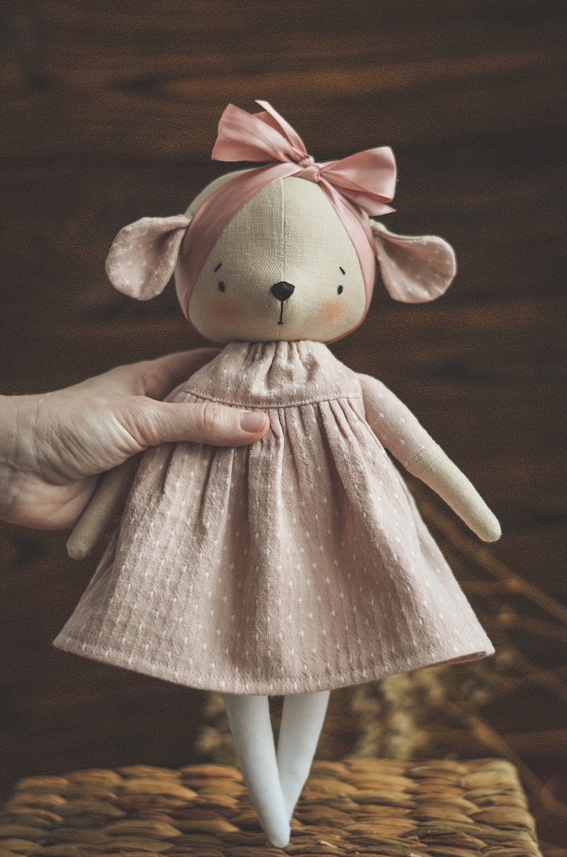 May include: A handmade doll with a pink dress and a pink bow in its hair. The doll has a light brown face with black eyes and a black nose. The doll is standing on a woven basket.