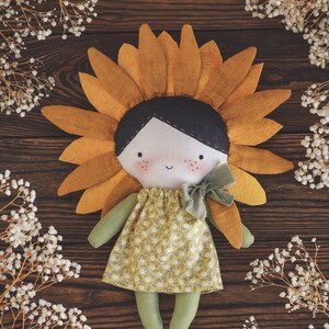 K&ouml;nnte beinhalten: Eine handgemachte Puppe mit einem Sonnenblumenkopf, die ein gr&uuml;nes Kleid mit Blumenmuster und eine gr&uuml;ne Schleife tr&auml;gt. Die Puppe sitzt auf einer Holzoberfl&auml;che mit wei&szlig;en Blumen.