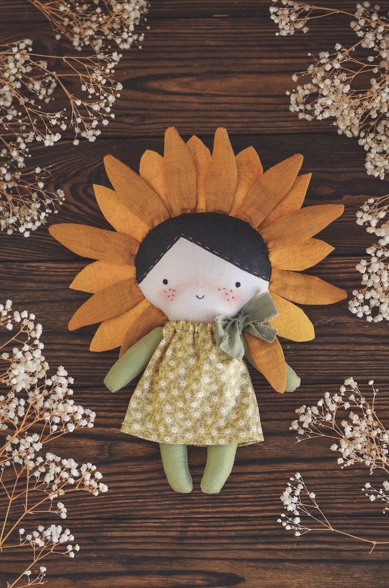 K&ouml;nnte beinhalten: Eine handgemachte Puppe mit einem Sonnenblumenkopf, die ein gr&uuml;nes Kleid mit Blumenmuster und eine gr&uuml;ne Schleife tr&auml;gt. Die Puppe sitzt auf einer Holzoberfl&auml;che mit wei&szlig;en Blumen.