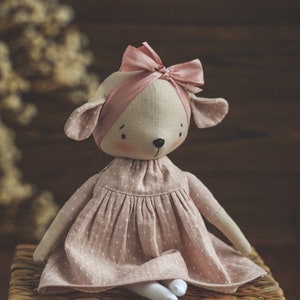 May include: A handmade plush toy in the shape of a deer wearing a pink dress and a pink bow. The toy is sitting on a woven basket.