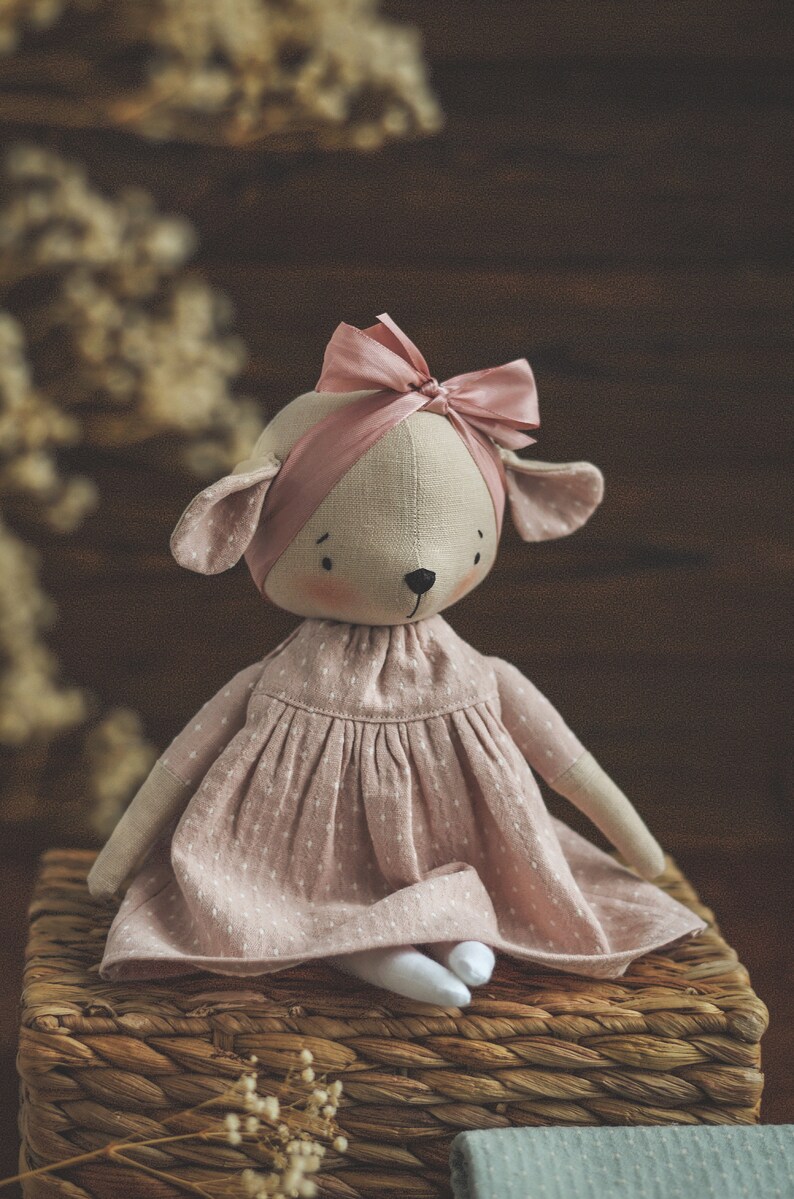 May include: A handmade plush toy in the shape of a deer wearing a pink dress and a pink bow. The toy is sitting on a woven basket.