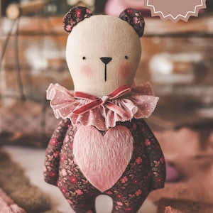 May include: A handmade plush teddy bear with a pink heart on its chest. The bear is wearing a pink ruffled collar and has a floral print fabric. The bear is sitting on a white surface with pink and brown lace.