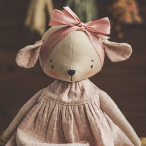 May include: A handmade doll with a pink dress and a pink bow on its head. The doll has a light brown fabric body and a white fabric face with black eyes and a black nose. The doll is sitting on a woven basket.