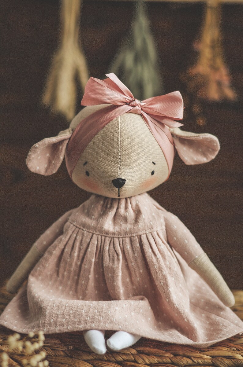 May include: A handmade doll with a pink dress and a pink bow on its head. The doll has a light brown fabric body and a white fabric face with black eyes and a black nose. The doll is sitting on a woven basket.