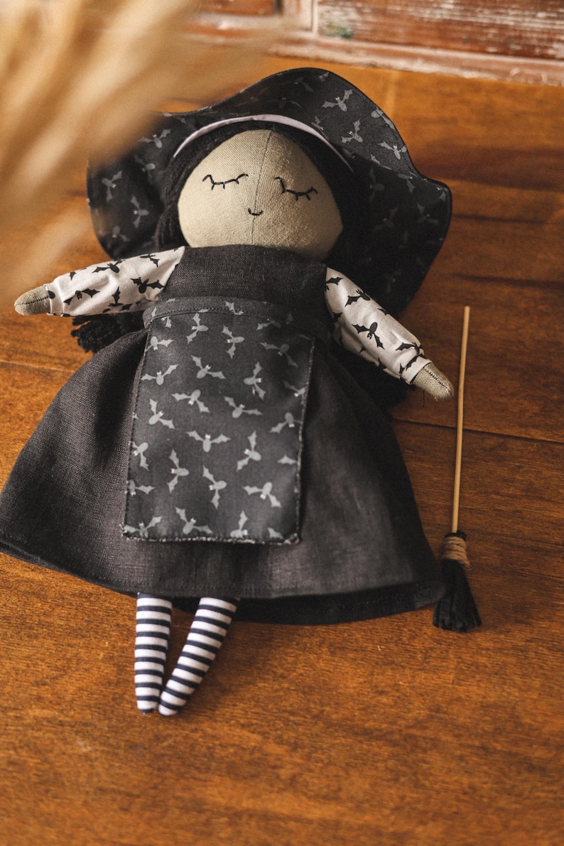 May include: A black witch doll with a black hat, a black dress with a white bat pattern, and striped stockings. The doll is holding a brown stick with a black broom at the end.