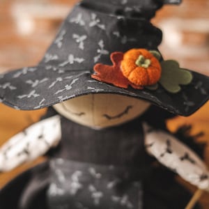 May include: A black witch doll with a black hat with a white bat pattern and a felt orange pumpkin and green leaf accent. The doll is wearing a black dress with a white bat pattern.