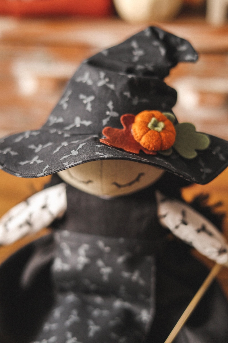 May include: A black witch doll with a black hat with a white bat pattern and a felt orange pumpkin and green leaf accent. The doll is wearing a black dress with a white bat pattern.