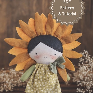 K&ouml;nnte beinhalten: Eine handgefertigte Puppe mit einem Sonnenblumenkopf, die ein gr&uuml;nes Kleid mit wei&szlig;em Blumenmuster und einer gr&uuml;nen Schleife tr&auml;gt. Die Puppe steht auf einer Holzoberfl&auml;che. PDF-Muster & Anleitung.