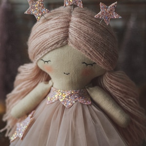 Op de afbeelding: Handgemaakte pop met een stoffen gezicht, gesloten ogen en blush. De pop heeft roze garen haar, een roze tule jurk en stervormige glitter accenten op het hoofd en de jurk. Een grillig, handgemaakt item.