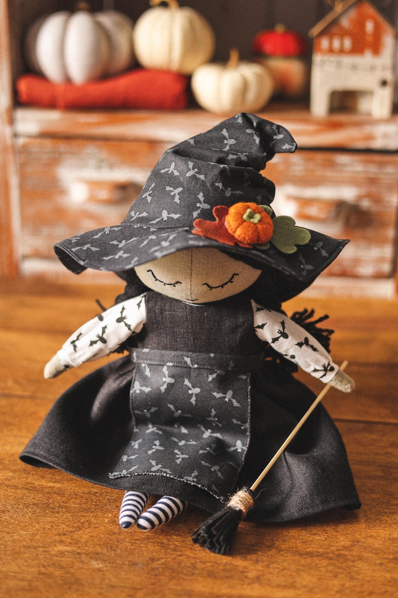 May include: A handmade witch doll wearing a black witch hat with a gray bat pattern, a black dress with a gray bat pattern, and black and white striped socks. The doll is holding a brown broomstick.
