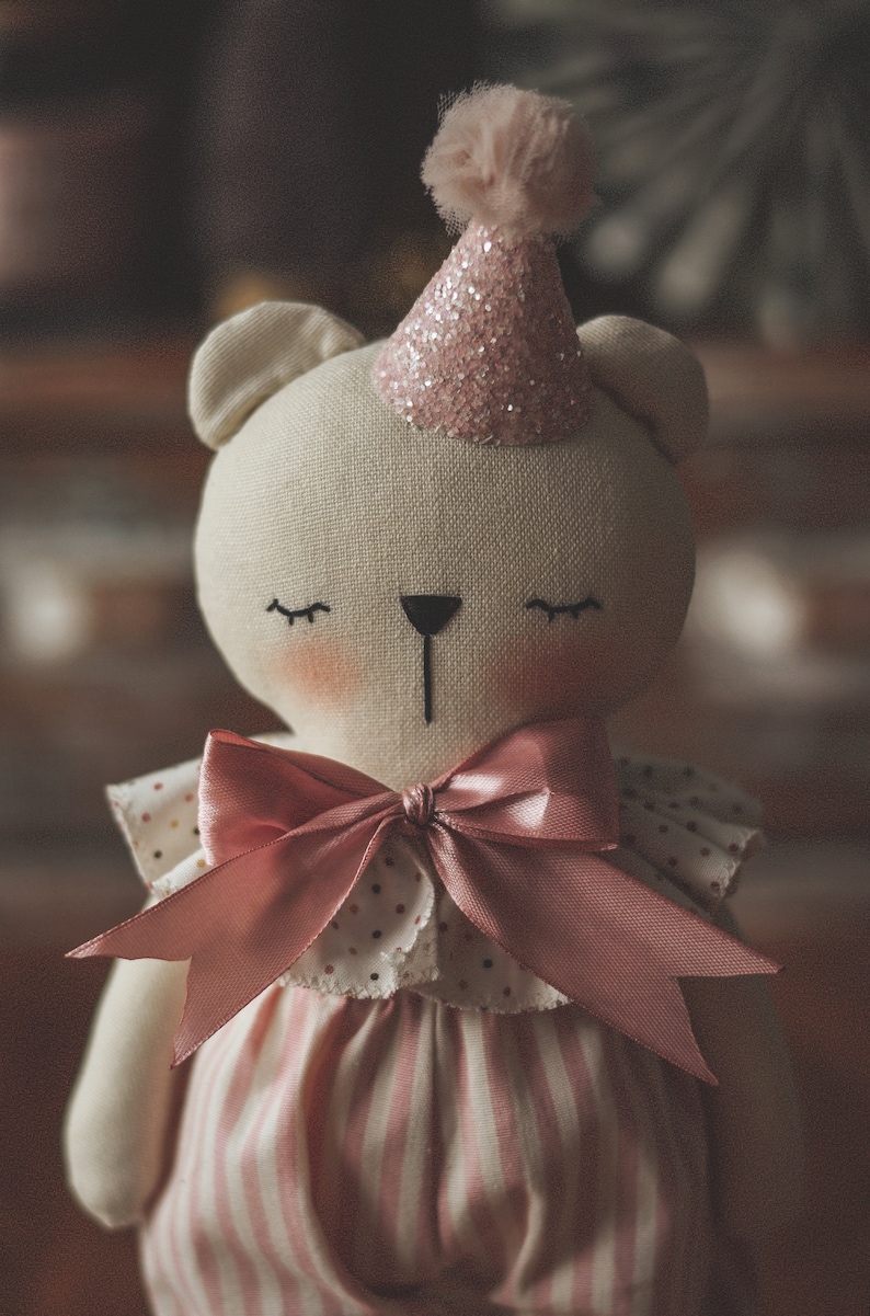 Op de afbeelding: Een witte knuffelbeer die een roze-wit gestreept jurkje draagt en een roze glitter-partyhoed met een witte pompon bovenop. De beer heeft een roze strik om zijn nek en gesloten ogen.