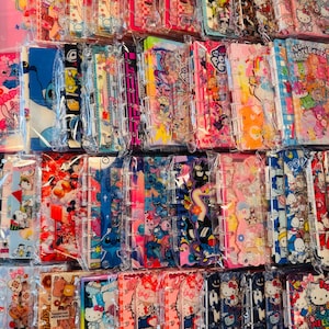 May include: A collection of colorful phone cases featuring various cartoon characters, including Hello Kitty, Stitch, and Mickey Mouse. The cases are in clear plastic packaging, with designs in pink, blue, and other bright colors.