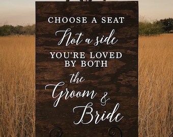 May include: A rustic wooden sign with the text "Choose a seat, not a side. You're loved by both the groom & bride." The sign is standing on a black easel in a field of tall grass.