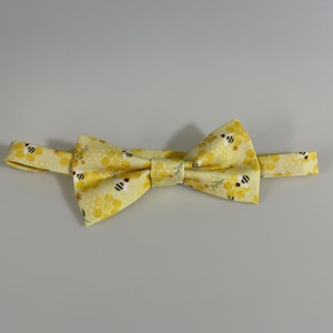 Könnte beinhalten: Gelbe Fliege mit einem sich wiederholenden Muster aus Bienen und Waben. Die Fliege ist aus Stoff gefertigt und hat einen verstellbaren Riemen. Ein formelles Modeaccessoire.