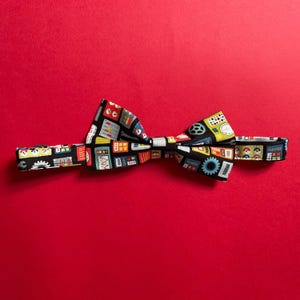May include: A black bow tie with a colourful pattern of various electronic components and gears. The bow tie is on a red background.
