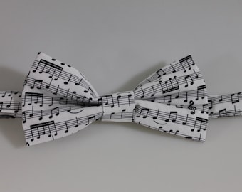 Puede incluir: Una pajarita preatada con fondo blanco y un estampado de pentagramas y notas musicales en negro. La tela muestra varios símbolos musicales, incluyendo claves de sol y notas dispuestas sobre líneas horizontales en toda la superficie.