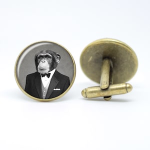 May include: A pair of cufflinks featuring a black and white image of a chimpanzee wearing a tuxedo and bow tie. The cufflinks are made of metal with a bronze finish.