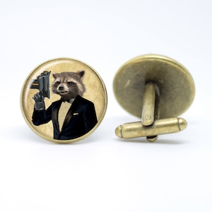 May include: A pair of cufflinks featuring a round glass cameo with a cartoon image of a raccoon wearing a tuxedo and holding a gun. The cufflinks are made of bronze metal.